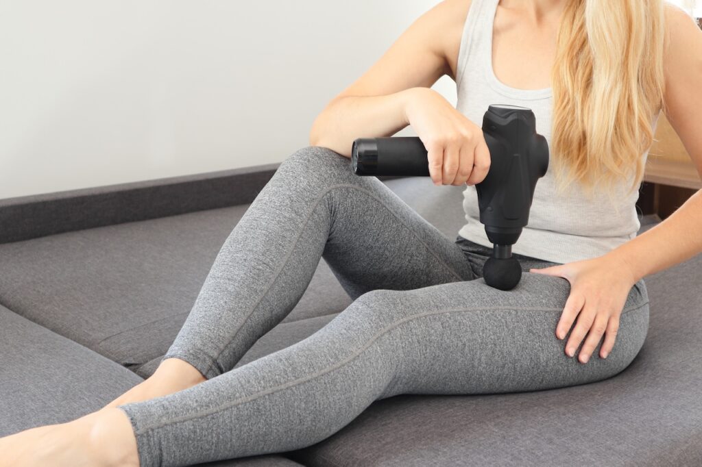 self massage of women's legs with a perfecting gun at home. athletic woman applying therapeutic percussive massage gun sitting on sofa at home.