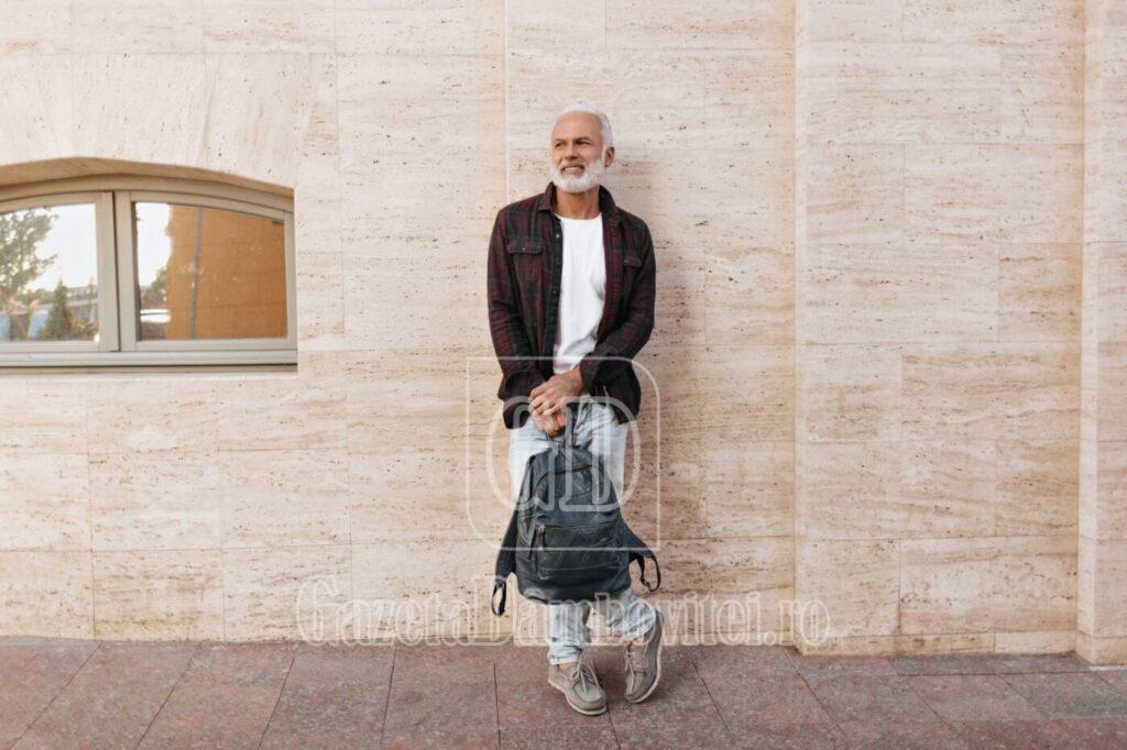 man in jeans and shirt leans on wall and holds backpack. gray haired adult fashionable guy in moder